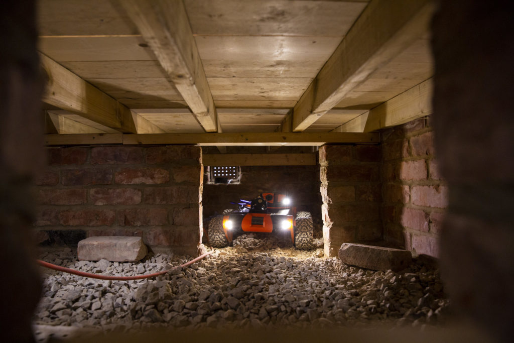 Small robot with lights maneuvers brickwork in the crawl space of a home to spray insulation.