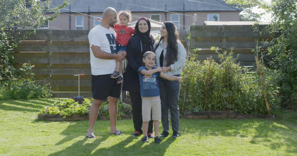 A family of five stand in a garden. A father holds a young child on his hip and the mother stands between him and teen daughter. Another young child stands in front of her.