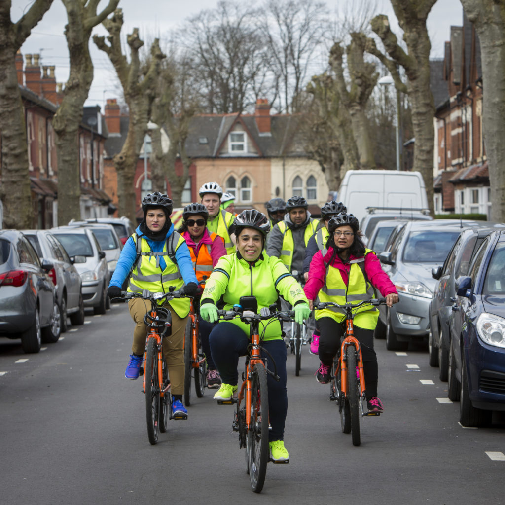 A group of people in multicoloured clothing and hi-vis jackets cycle down a street. 