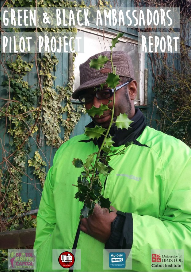 A black man in a green hi-vis jacket and a cap holds a long branch of spiked leaves that partially covers his face. 