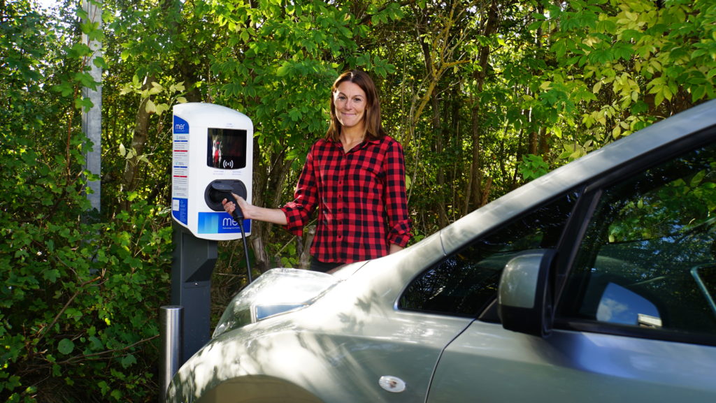Woman plugs electric vehicle into a charging station in front of forest.