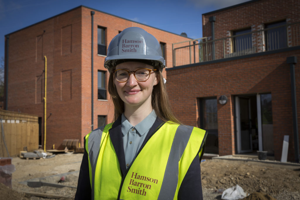 A young white woman wearing glasses, a hardhat and yellow hi-vis jacket stands in front of a building site. 