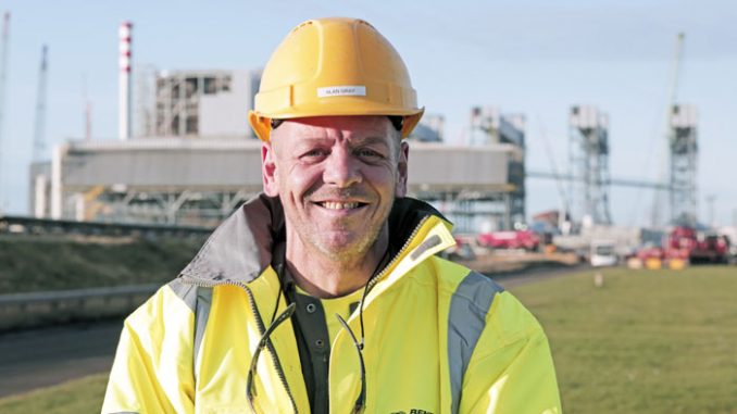 A man in a hardhat and yellow hi-vis jacket smiles for the camera. A biomass station is visible in the background. 