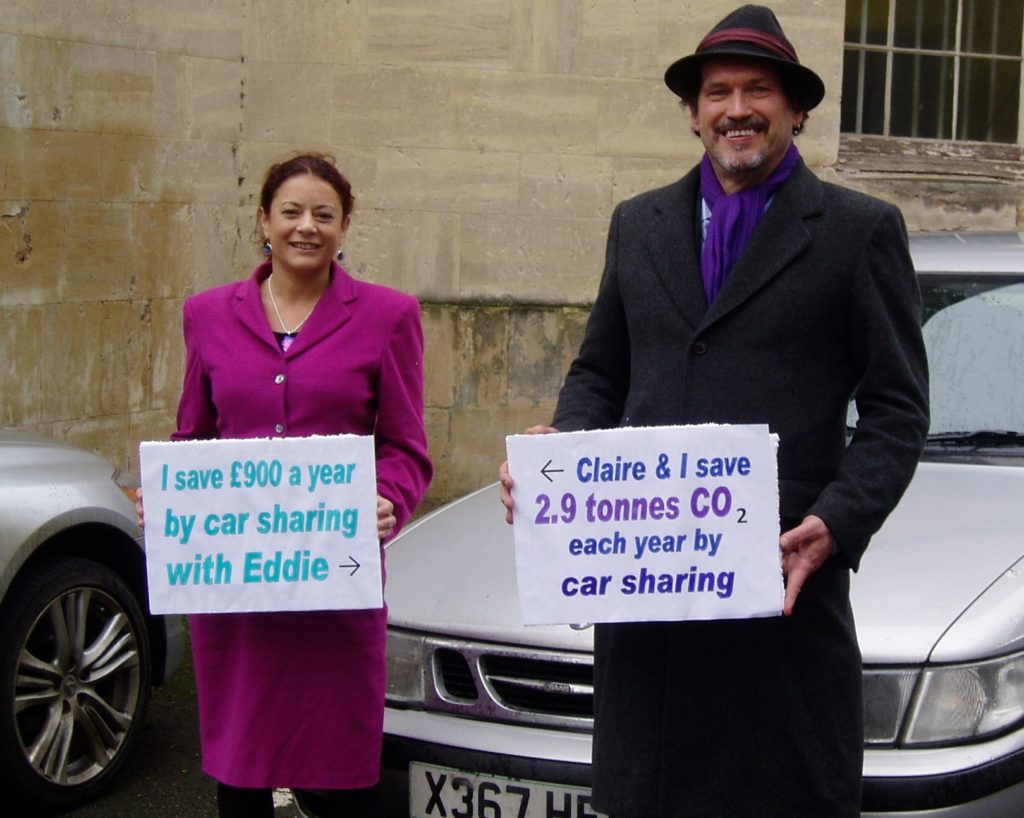 Two people stand next to each other in front of a silver car. A woman in a pink dress holds a sign saying 'I save £900 a year by car sharing with Eddie'. A man holds a sign saying 'Claire & I save 2.9 tonnes CO2 each year by car sharing'. 