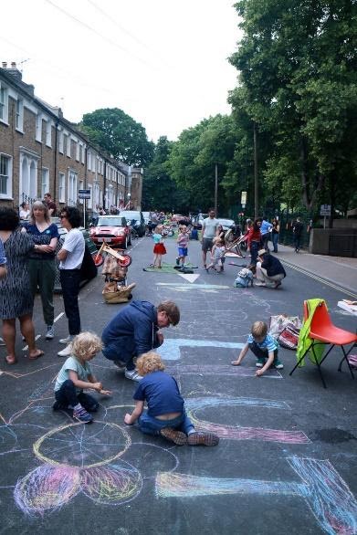 A shot of a busy, mostly car-free street: parents chat, children do chalk drawings on the road and play games. 