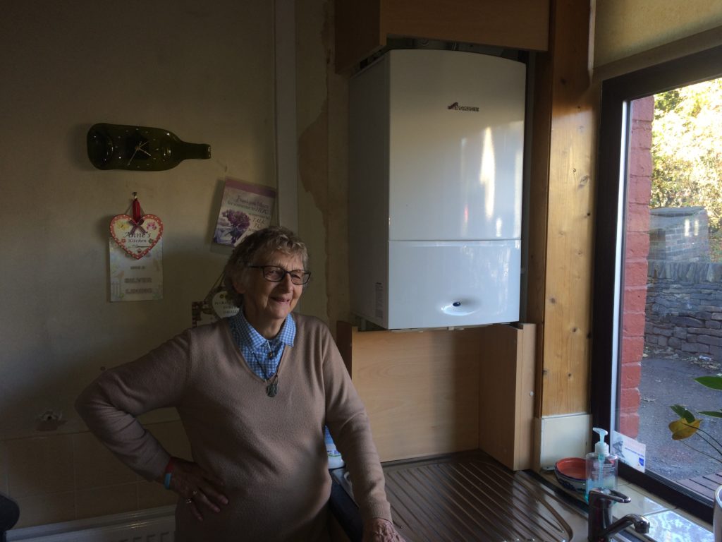 An elderly individual stands in their kitchen next to a boiler. 
