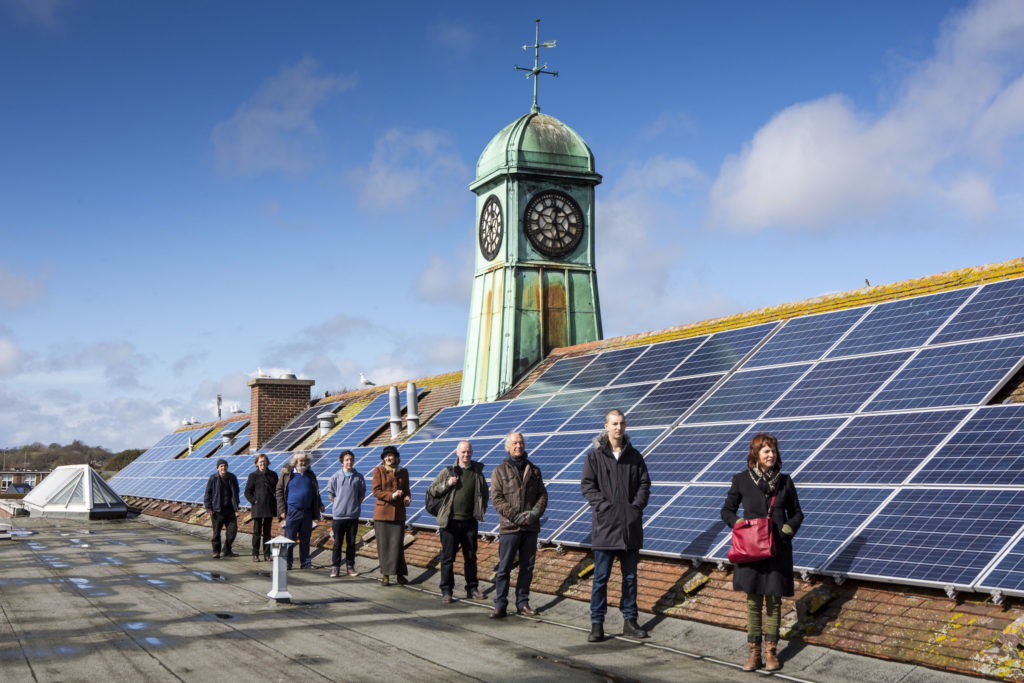 A line of people stand on the roof of a school building next to a long row of solar PV panels. 