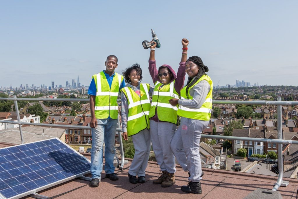 A group of four young black people stand on a roof next to a solar panel. Each wears a yellow hi-vis jacket. One young woman has her hands raised in the air - she is also holding a drill. 