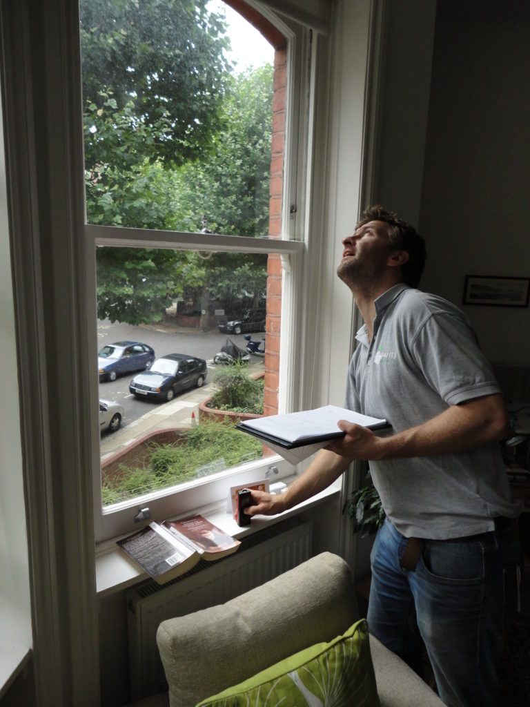 A man inside a house by a window, looking upwards. He holds a clipboard. 