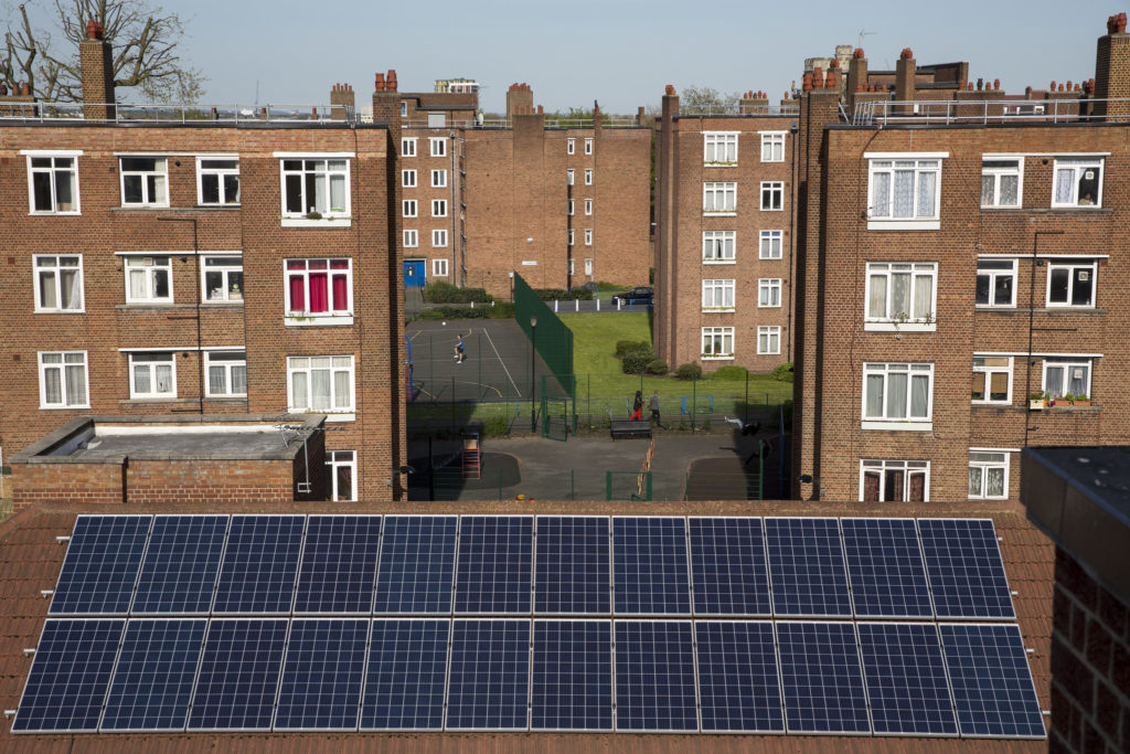 A shot of solar panels installed on a roof overlooking a residential area in Hackney comprised of blocks of flats. 