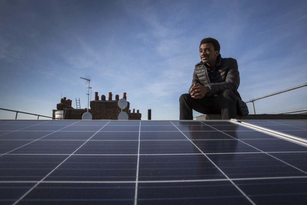 A young black man sits next to a rooftop solar panel with a background of blue sky and chimneys. 