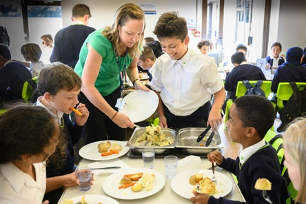 A female teacher helps serve lunch to four smiling students eating in the cafeteria.
