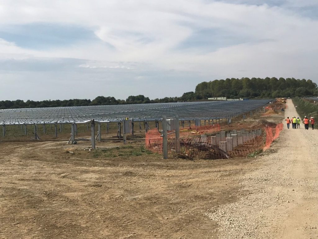 Expansive solar farm extends over a sandy clearing, workers in high-vis vests and hardhats walk next to it along a dirt path.