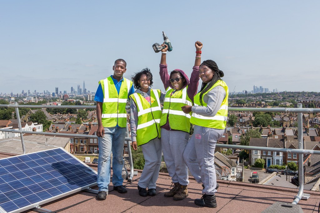 4 individuals in hi-vis vests smile while standing on a city roof next to a solar panel - one raises arms in celebration and holds a drill.