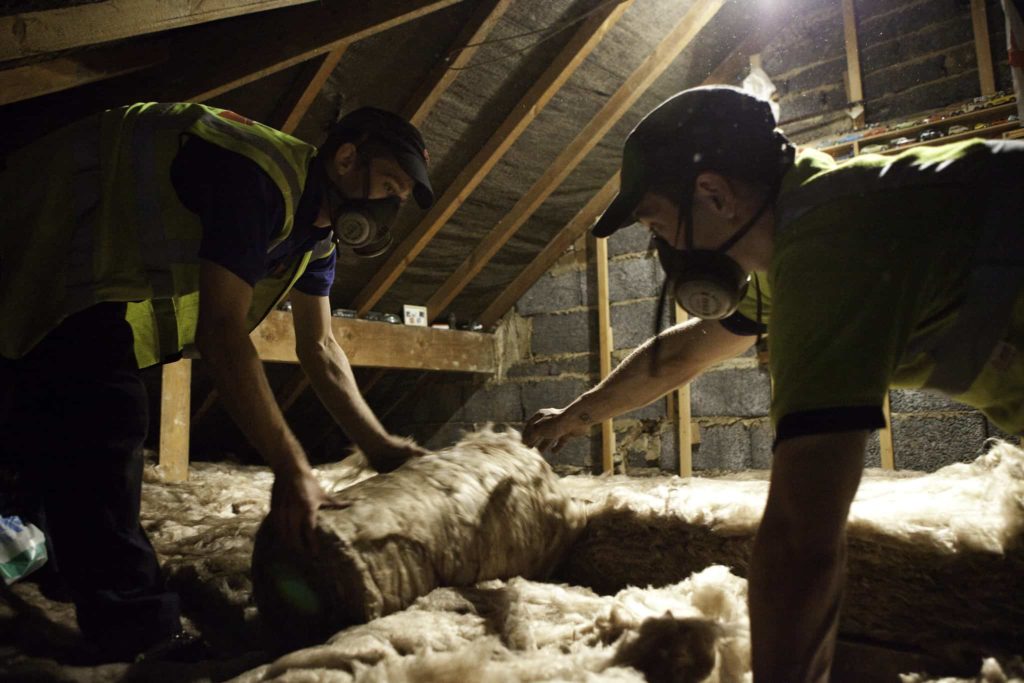 Two men wearing in dust masks and high-vis vests lay insulation in an attic.
