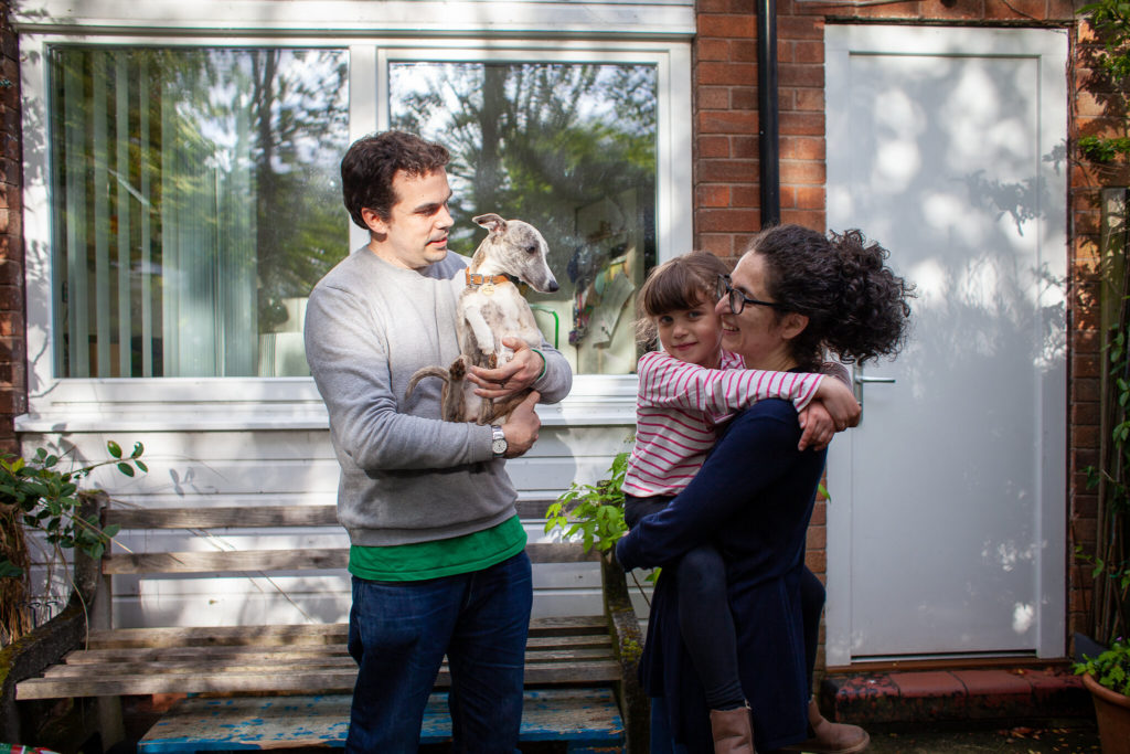 Man (left) holding small dog, looking toward smiling woman (right) holding and hugging little girl, standing outside of home.